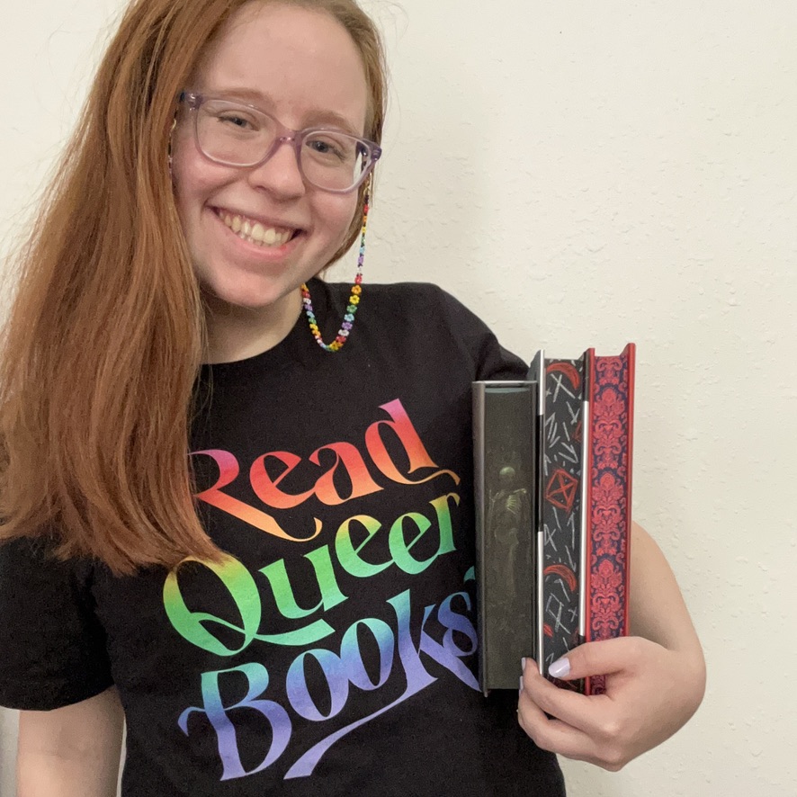 Haley, a pale person with long, straight auburn hair past their shoulders, stands in front of a cream-colored wall, smiling at the camera. They are shown from the torso up, wearing a black shirt with "Read Queer Books" in rainbow lettering. They are wearing light purple glasses with a rainbow flower glasses chain that drapes on their shoulder. In their arm, they are holding 3 hardcover books with colorful sprayed/painted edges (green, black/red/white, and red floral).